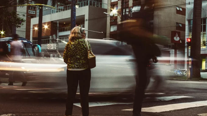 a woman standing at a crosswalk in a downtown city with streaky cars and people racing by
