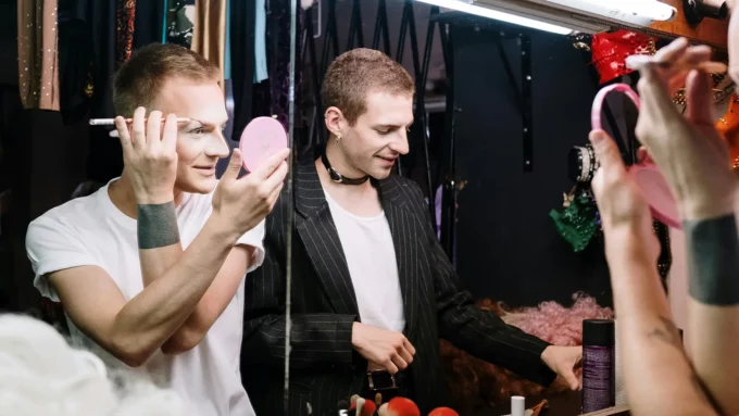 two people presenting as male putting on makeup in a mirror