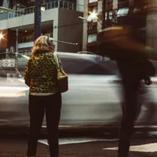 a woman standing at a crosswalk in a downtown city with streaky cars and people racing by