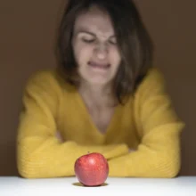 a white woman sitting at a table with a disgusted look at apple she does not like