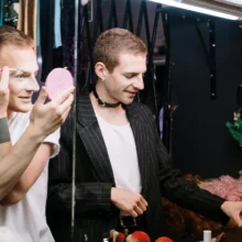 two people presenting as male putting on makeup in a mirror