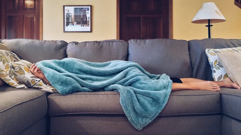man lying on couch with blanket over head sick
